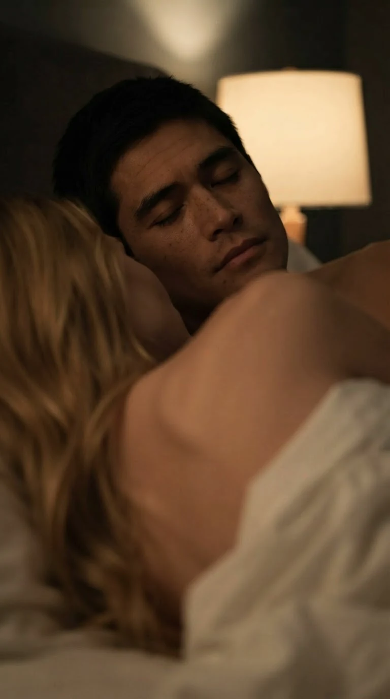 A man lying still with his eyes closed in a dim room, a blonde woman's bare shoulder beside him.