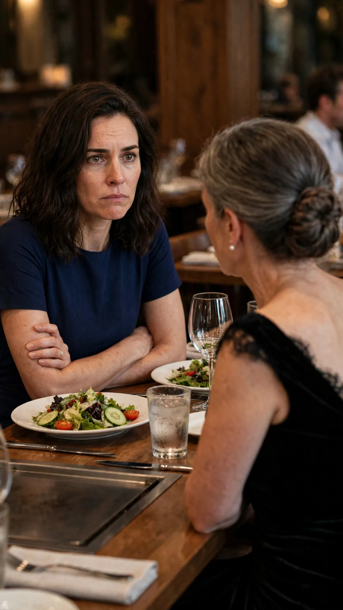 Lena sits alone at the far end of the table, a woman across from her looking away.